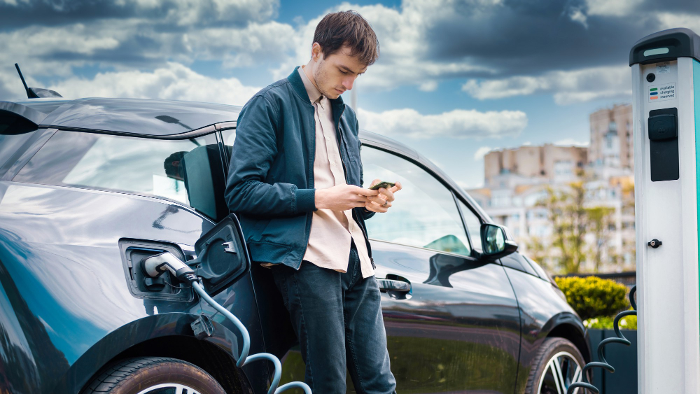 Young man checking phone while charging electric car at EV station