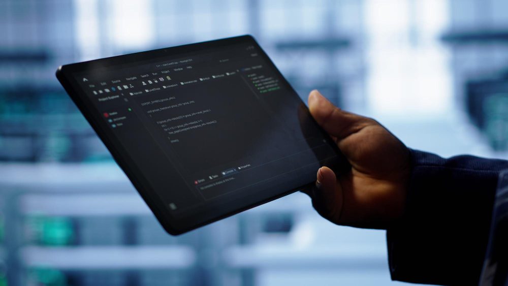 Hand holding tablet displaying software code and AI development interface in a modern data center environment